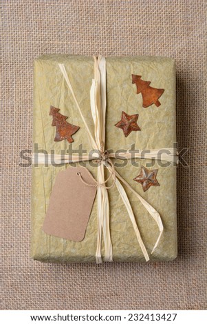 High angle closeup of a gold tissue paper wrapped Christmas present on a burlap surface. The gift is tied with raffia and has antique star and tree tin ornaments. Vertical format.