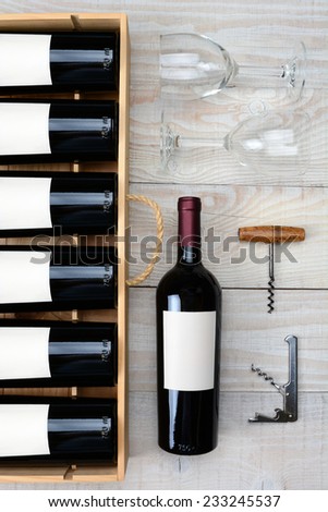 High angle shot of a case of red wine bottles next to a single bottle two wineglasses and cork screws on a rustic white wood table. Vertical format.