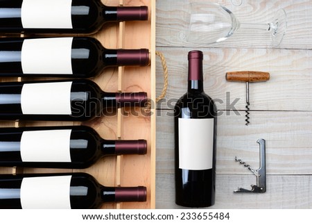 High angle shot of a case of red wine bottles with blank labels next to a single bottle a wineglass and cork screws on a rustic white wood table. Horizontal format.