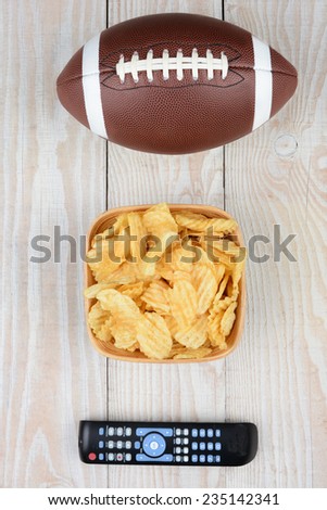 Getting ready to watch a Bowl Game. High angle shot of an American football, a bowl of potato chips and a TV remote on a white wood table. Vertical format