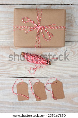 High angle shot of a Christmas Package wrapped in plain brown paper and tied with red and white string. Three blank tags and spool of string are below the gift on a white rustic table.