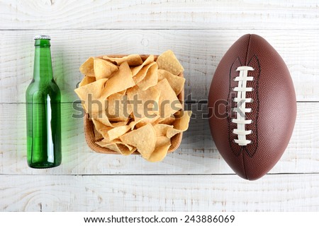 Beer bottle, bowl of chips and an American style football on a rustic whitewashed wood surface. Horizontal format. The bottle is without label.