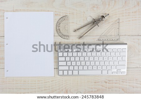 High angle shot of a computer keyboard next to a sheet of graph paper, a compass and protractor, on a whitewashed wood table, in a home office.