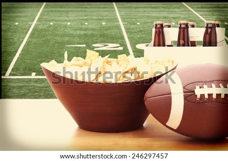 Chips, football and Six Pack of Beer on a table in front of a big screen TV with a Football field. Great for Super Bowl themed projects. Horizontal format with instagram effect applied.