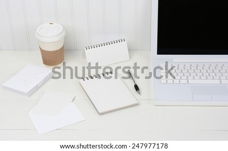 Closeup of a neat white desk with Laptop, disposable coffee cup, note pad, envelopes and pen. A wood desk with a beadboard wall behind. The image is primarily shades of white.