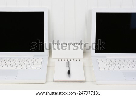 Closeup of two laptops in a white work station with pad and pen in the middle. The computers are against a beadboard wall with blank screens. Horizontal format.