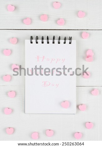 Happy Valentine\'s Day on a note pad in pink type. Shot from a high angle on a white wood table. The pad is surrounded by pink candy hearts in a random pattern.