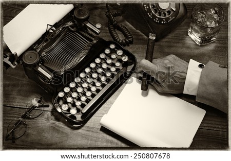 Closeup of a writer at his desk with a typewriter, rotary telephone, glass of whiskey and a cigar. Black and white toned image for a vintage feel. Only the mans hand holding a cigar is shown.