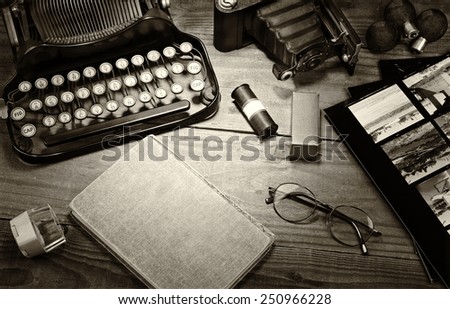 Closeup of a vintage photography still life with typewriter, folding camera, loupe, roll film, flash bulbs, contact prints and book on a wood table. Black and white toned image for a vintage feel.