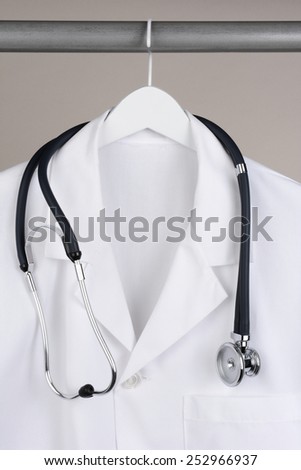 Closeup of a doctor\'s lab coat and stethoscope on hanger against a neutral background. White coat on a white hanger with a gray background in vertical format.