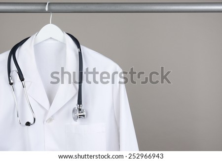 A doctor\'s lab coat and stethoscope on hanger against a gray background. Closeup on a white hanger with a gray background in horizontal format with copy space.