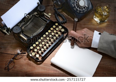 Closeup of a writer at his desk with a typewriter, rotary telephone, glass of whiskey and a cigar. A vintage feel with only the mans hand holding a cigar being shown.