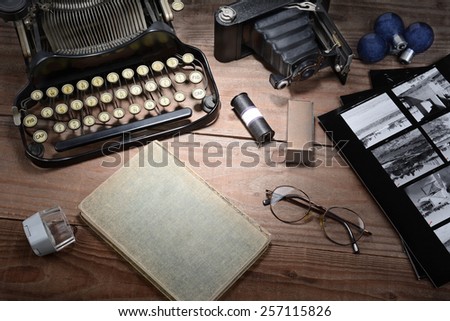 Closeup of a retro photography still life with typewriter, folding camera, loupe, roll film, flash bulbs, contact prints and book on a wood table. Black and white toned image for a vintage feel.