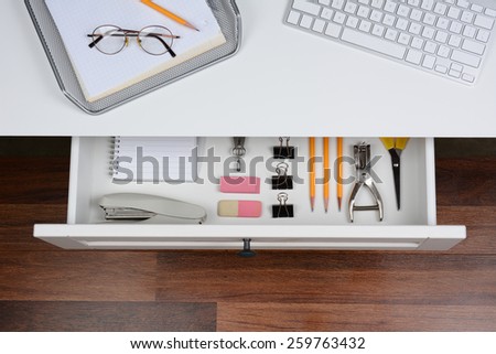 High angle shot of an open desk drawer showing the items inside. The top of the desk has a computer keyboard and wire in-box with paper and pencil. The drawer has pencils, erasers, stapler and more.