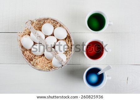 High angle shot of a basket full of eggs to be dyed for Easter. Three cups of dye with eggs soaking are next to the baskets of un-dyed eggs on a white wood kitchen table.