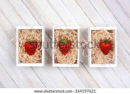 Three mini wood crates filled with straw each with a single large ripe strawberry. High angle shot on a rustic whitewashed wood table with copy space.