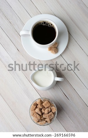 High angle shot of a cup of coffee, cream pitcher, and a bowl of natural sugar cubes. Vertical format on a rustic whitewashed table.