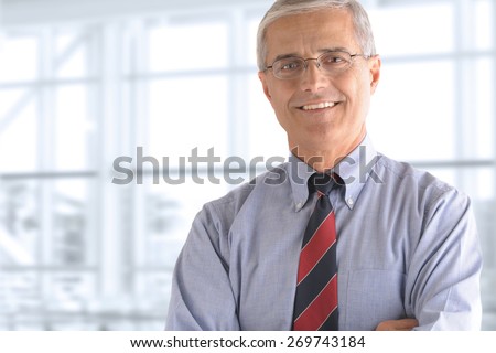 Portrait of a mature businessman standing in front of a large office window. The man is smiling and has his arms crossed.