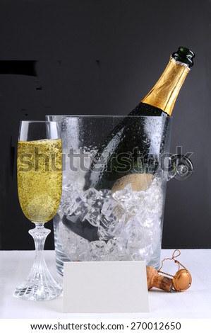 Closeup of a crystal champagne ice bucket and a full glass of bubbly. A blank table tent and cork in front of the open bottle lay on the white table cloth, over a light to dark background.