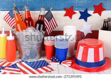 Fourth of July still life. A picnic table with a bucket of soda, flags, Uncle Sam hat, 6 pack, ketchup and mustard with a red, white and blue wall behind.