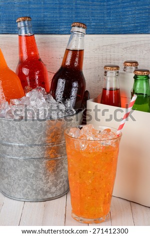 An ice bucket full of soda bottles, a six pack carrier, and a glass of orange soda with straw. Closeup on a rustic set of white washed wood and one blue board.