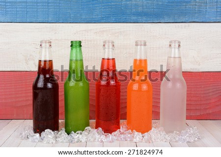 Five different bottles of soda on a rustic wood picnic table with a red white and blue background.