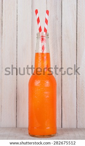 Closeup of a bottle of orange soda with two red and white striped drinking straws. Vertical format on a rustic wood background. The bottle is covered with condensation.