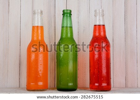 Three soda bottles on a wood background. The bottles are open and covered with condensation. Orange soda, Strawberry soda and Lemon Lime are represented.
