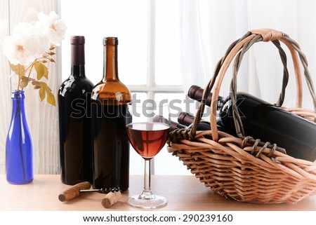 Wine still life with a basket of bottles and a glass of red wine and white roses in a vase on a table in front of a window with curtains. Horizontal format with vignette filter effect.