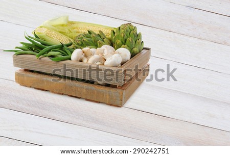 A wood crate fill with assorted vegetables on a rustic wood table. Mushrooms, Corn, Artichokes and Green Beans fill the box.