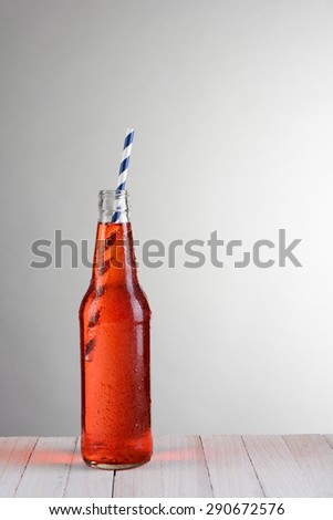 A single bottle of strawberry soda on a wood table against a light to dark gray background. The bottle has a blue and white drinking straw at an angle.