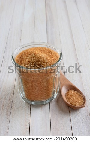 High angle shot of a glass bowl full of granulated brown sugar on a rustic whitewashed wood table. A wood spoon is laying next to the bowl.