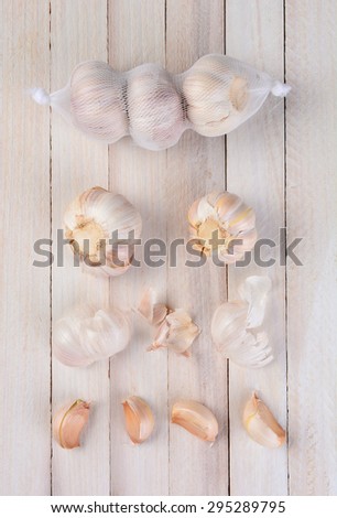 Overhead view of garlic on a rustic white table. Three bulbs in a net bag, two bulbs below the bag, scattered skin pieces and four clove segments.