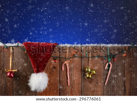 A rustic fence with christmas lights, bells, candy canes and a Santa Claus hat. Horizontal format with a light to dark blue night sky and snow effect.