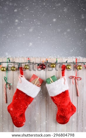 Two Christmas Stockings hanging on a rustic white fence with lights, jingle bells, and candy canes. Vertical format with snow effect.
