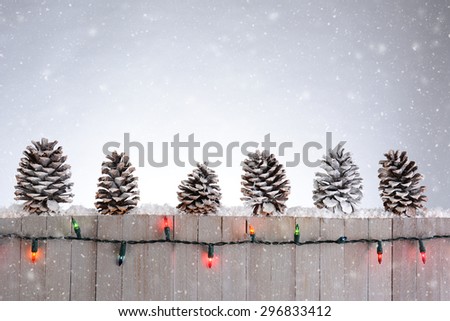 A rustic wood fence with pine cones lined up on top. Christmas lights and snow flakes round out the holiday scene.