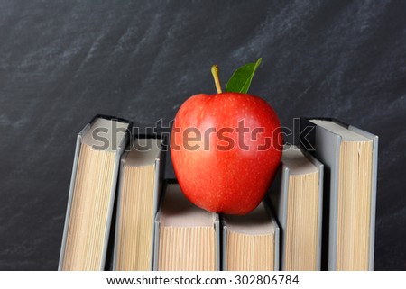 A red apple perched atop text books on a teachers desk with an out of focus chalkboard behind.