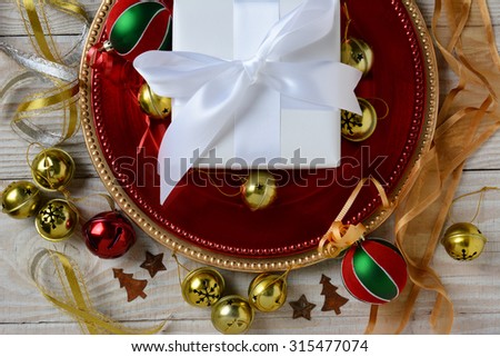 Overhead view of a white paper wrapped Christmas Present on red and gold chargers surrounded by holiday ornaments and ribbon. Horizontal format.