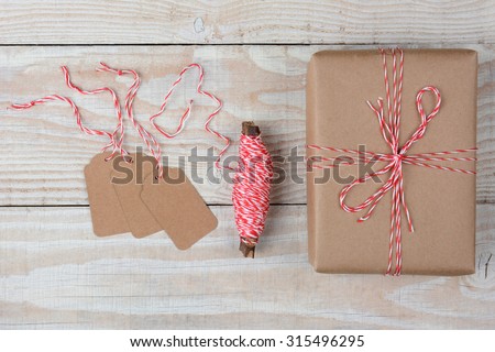 Overhead view of blank gift tags a spool of string and a plain brown paper wrapped present on a rustic whitewashed wood table.
