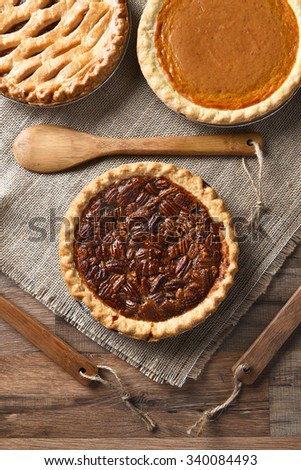 High angle vertical view of three pies for A thanksgiving feast. Pecan in the front with Apple and Pumpkin pies in the background. On burlap with wood utensils.