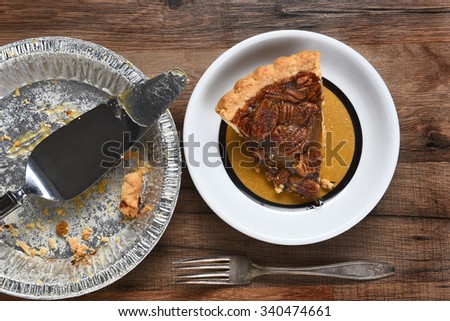 High angle view of a slice of pecan pie on a plate next to the empty pie tin with server. On a wood table with fork.