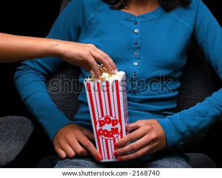 Teenage girl holding a popcorn bucket at the movies with a friend reaching into bucket for a handful.