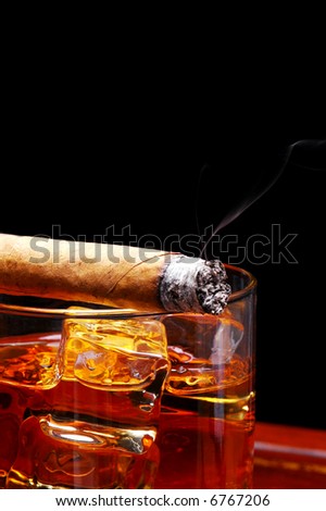 Lit Cigar resting on the rim of a glass of whiskey on the rocks with wisp of smoke over a black background with copy space.