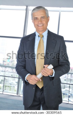 Smiling Middle Aged Businessman in dark suit standing and holding a rolled up newspaper in office setting