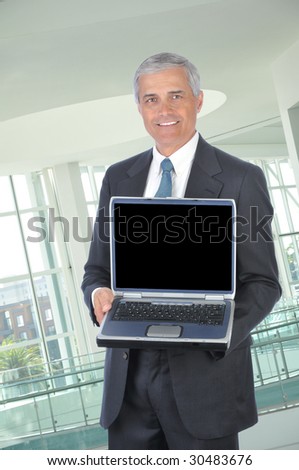 Smiling Middle Aged Businessman in dark suit standing with laptop computer open and facing the camera in office setting