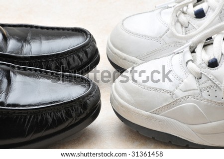 Closeup of two pairs of shoes, black slip-on loafers and white sneakers. The shoes are toe to toe showing only the front half of each shoe. Play shoes / work shoes concept. Horizontal format.