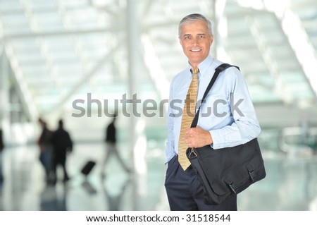 Smiling Middle Aged Businessman Standing with Travel Bag Over Shoulder in Airport Concourse - blurred travellers in background