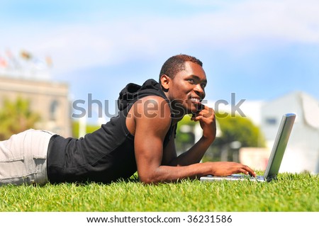 African-American male with laptop laying in the grass in park setting - blue sky and out of focus buildings in background. Horizontal composition - torso only.