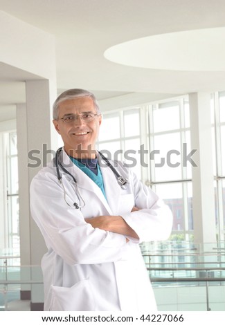 Middle aged doctor wearing lab coat and surgical scrubs with arms crossed in modern medical facility