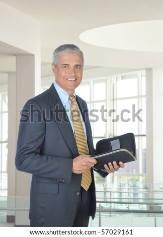 Smiling middle aged businessman holding a planner notebook. Man is standing in the atrium of a modern office building. Vertical format.